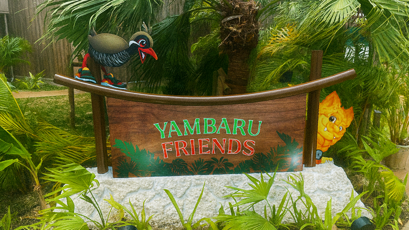 やんばるフレンズの木製看板。ヤンバルクイナのキャラクターが上に立ち、右側から黄色い獅子のようなキャラクターが顔をのぞかせている。背景は南国の植物で、雨に濡れてしっとりとした雰囲気。